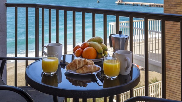 Een ontbijttafel met croissants, fruit, sinaasappelsap en koffie op een balkon met uitzicht op zee.