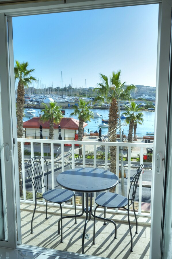 Un balcon avec une table et des chaises, donnant sur une marina où l’on peut admirer des bateaux et des palmiers.
