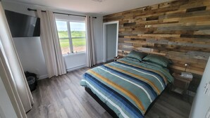 Ein Schlafzimmer mit großem Fenster, einem Bett mit gestreifter Bettdecke, einem Holz-Kopfteil und einem Nachttisch mit Lampe.