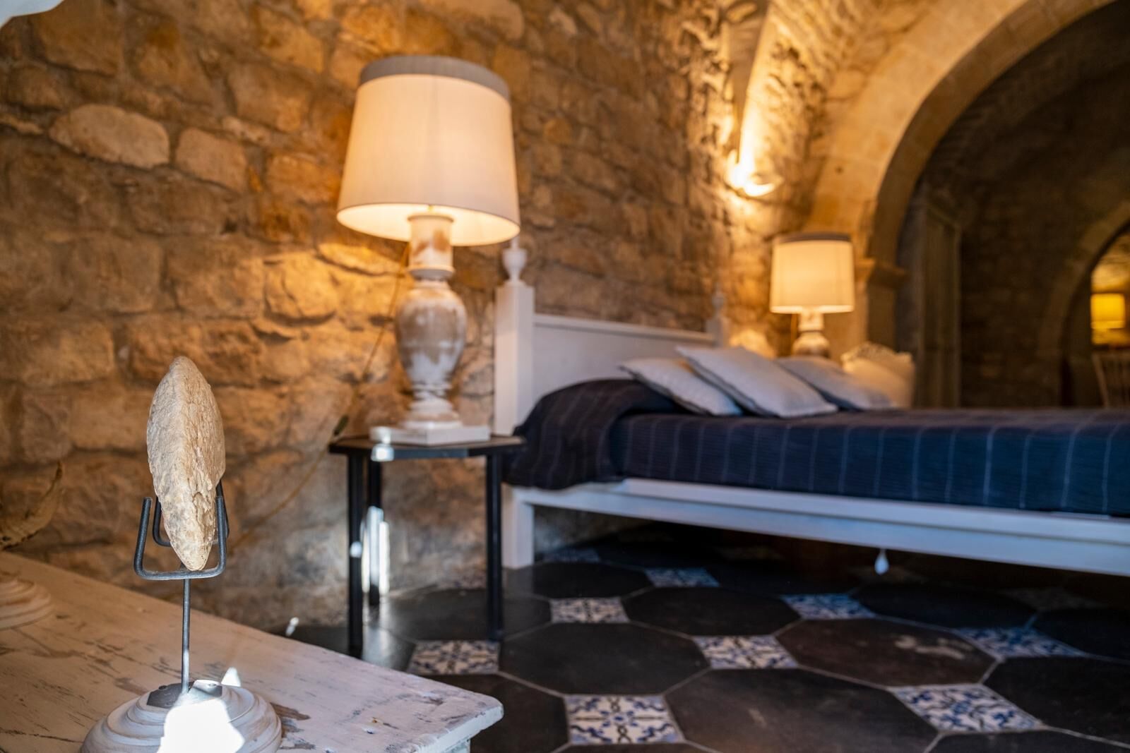 A bedroom with a stone wall, a bed with a dark blanket, a bedside table with a lamp, and a decorative stone on the floor.