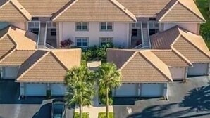 A multi-story building with a sloped roof, a parking area, and a view of a lake with dense greenery in the background. - Spacious 3-bedroom first floor coach home with attached garage in North Naples (Naples)