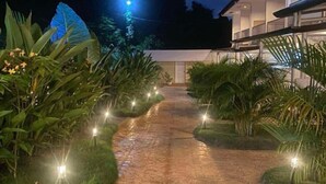 A paved pathway leading to a modern building with a glass facade, surrounded by lush greenery and illuminated by ground lights. - Tropical Comfort & Charm (Puerto Princesa)
