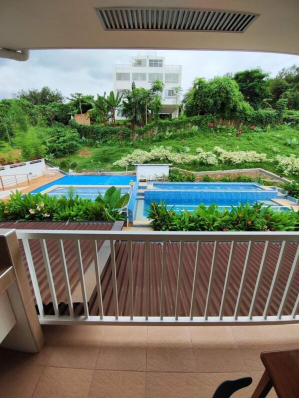 A balcony with a view of a swimming pool, surrounded by lush greenery and a multi-story building in the background.