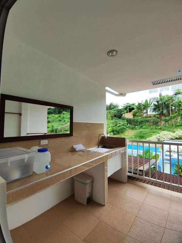 A modern bathroom with a large mirror, a sink, and a toilet. There is a small trash bin and a storage container on the counter. The bathroom has a view of a pool and greenery outside.