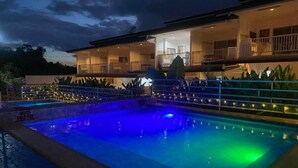 A swimming pool with a blue and green light feature, surrounded by a fence and a building with multiple balconies.