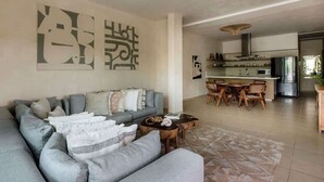 Una sala de estar moderna con un sofá seccional gris, un comedor con mesa y sillas de madera, y una cocina con electrodomésticos de acero inoxidable.