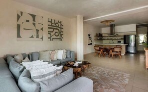 A modern living room with a grey sectional sofa, a dining area with a wooden table and chairs, and a kitchen with stainless steel appliances.