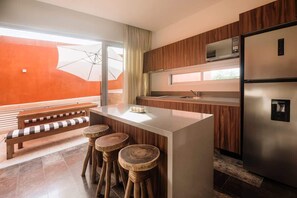 A modern kitchen with a central island, wooden bar stools, a bench, and a large window with an umbrella. - Anaya Luxury Jungle View Penthouse Rooftop Pool (Tulum)
