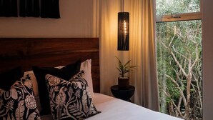 Quarto com cabeceira de madeira, duas almofadas estampadas, uma planta na mesa de cabeceira e vista para árvores pela janela.