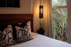 A bedroom with a wooden headboard, two patterned pillows, a bedside plant, and a view of trees through a window. - Anaya Luxury Jungle View Penthouse Rooftop Pool (Tulum)