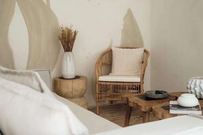 Ein Zimmer mit einem Korbstuhl, einer weißen Vase mit Trockenblumen, einem runden Holztisch und einem weißen Kissen.