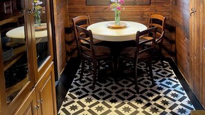 A wooden cabin interior with a round dining table, chairs, a window with a hanging light, and a patterned floor.