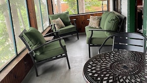 A covered patio with wooden ceiling and large windows, furnished with green chairs and a metal table.