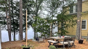 An outdoor patio with a wooden table and chairs, surrounded by trees and a view of a body of water.