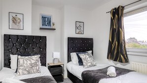 A hotel room with two beds, each with a black headboard and floral-patterned pillows. There are framed artworks on the walls, a small bedside table with a lamp, and a window with curtains. - Last Min Weekend Stay, Trafford, Pets Welcome (Sale)