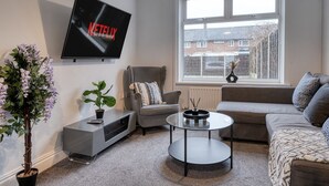 A modern living room with a grey sofa, a grey armchair, a round coffee table, a TV mounted on the wall displaying 'NETFLIX', and a window with a view of houses. - Last Min Weekend Stay, Trafford, Pets Welcome (Sale)