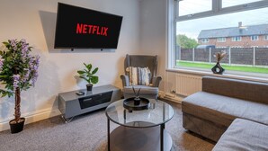 A modern living room with a flat-screen TV displaying 'NETFLIX', a grey armchair, a grey sofa, a glass coffee table, and a potted plant. - Last Min Weekend Stay, Trafford, Pets Welcome (Sale)