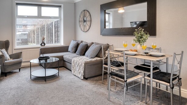 A modern living room with a grey sofa, a round coffee table, and a dining table set for two. A large mirror and a wall clock are visible. - Last Min Weekend Stay, Trafford, Pets Welcome (Sale)