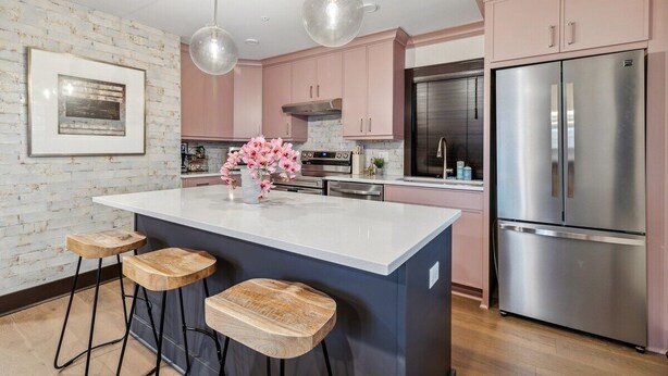 Uma cozinha moderna com uma ilha central, banquetas, armários cor-de-rosa, eletrodomésticos de aço inoxidável e iluminação pendente.