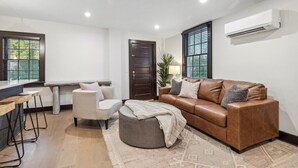 Uma sala de estar moderna com um sofá de couro marrom, um banquinho redondo, uma mesinha e um banqueta. Há uma porta, uma janela com persianas e um aparelho de ar condicionado na parede.
