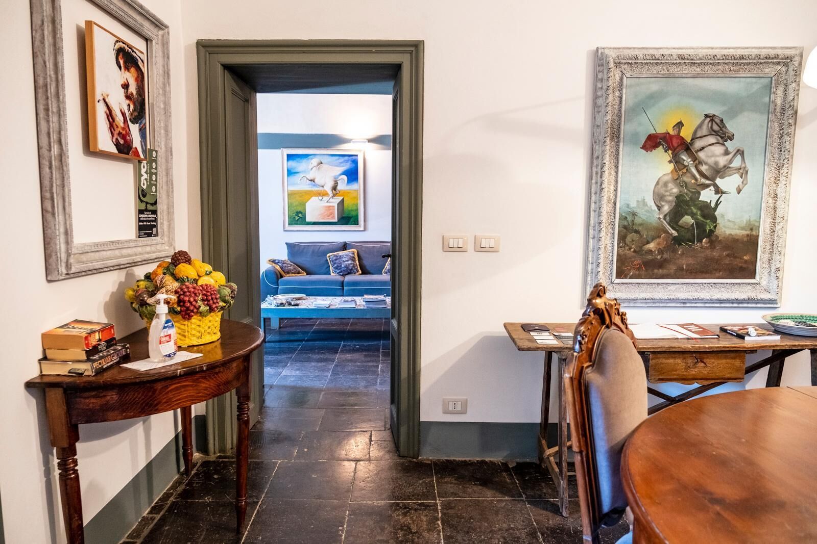 Un coin repas avec une table en bois, des chaises et un vase rempli de fruits. Un tableau orne le mur et un canapé se trouve au fond de la pièce.