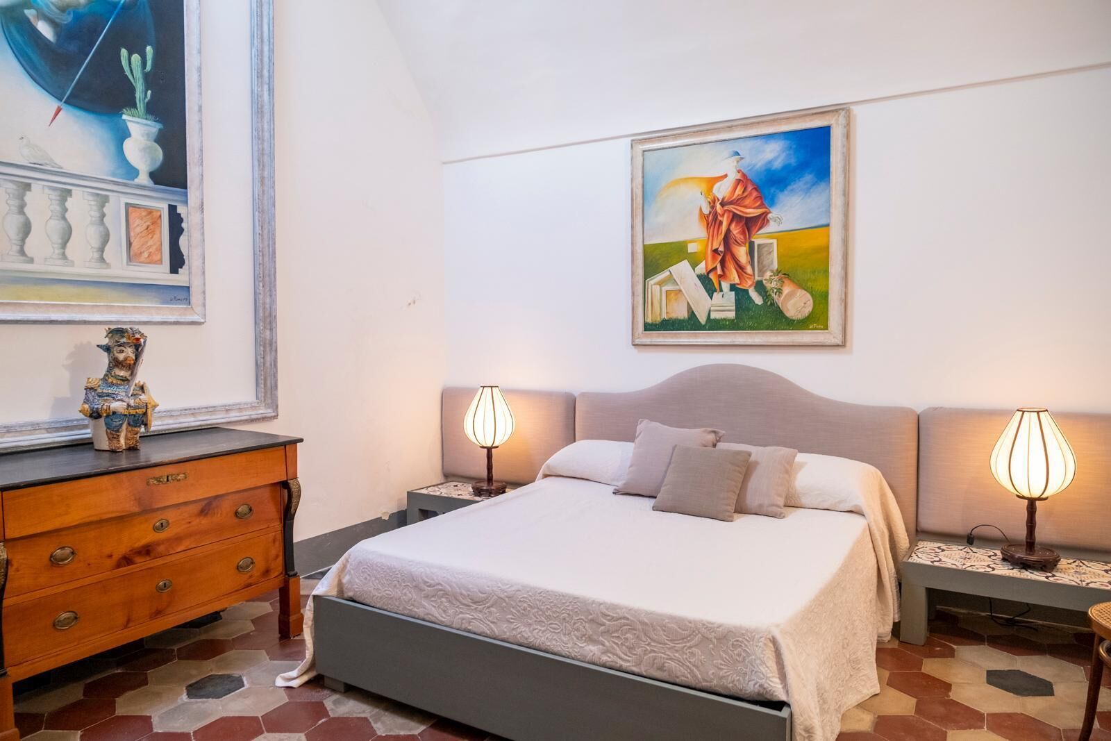 A bedroom with a bed, two bedside tables with lamps, a dresser, a painting on the wall, and a decorative item on the dresser.