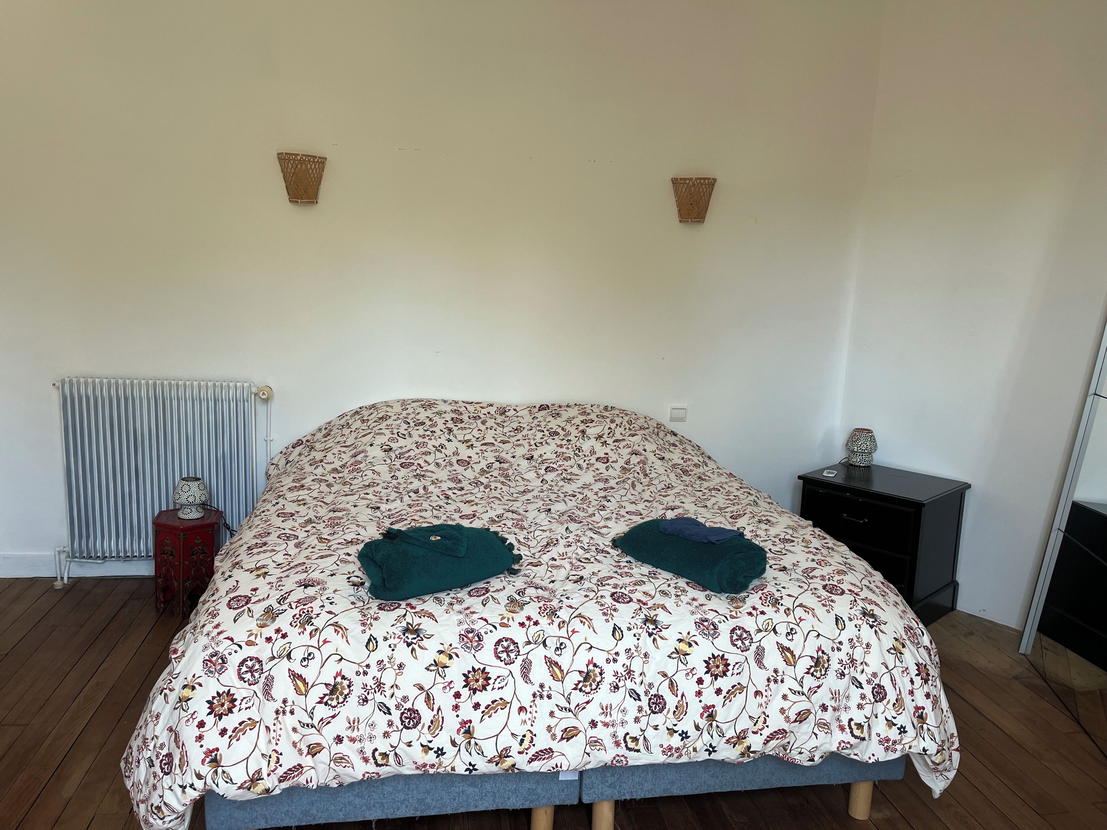 Une chambre à coucher avec une couvre-lit à motifs floraux, deux oreillers, un radiateur, une petite table avec une lampe et une table de chevet avec une lampe.