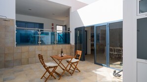 Pátio iluminado pelo sol, com mesa e cadeiras, guarda-corpo de vidro e vista para o interior.