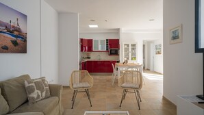 Uma sala moderna com um sofá, duas cadeiras de vime, uma mesa de centro e vista para uma cozinha com armários vermelhos e uma área de jantar.