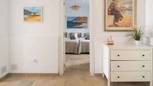 Quarto de hotel com uma cama, uma pequena mesa de trabalho e uma planta em vaso. Há quadros nas paredes e vista para a praia através da porta.