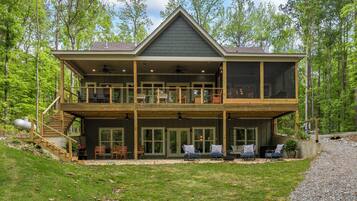 Une maison en bois à deux étages, dotée d’un balcon, de grandes fenêtres et d’un espace extérieur aménagé et abrité.