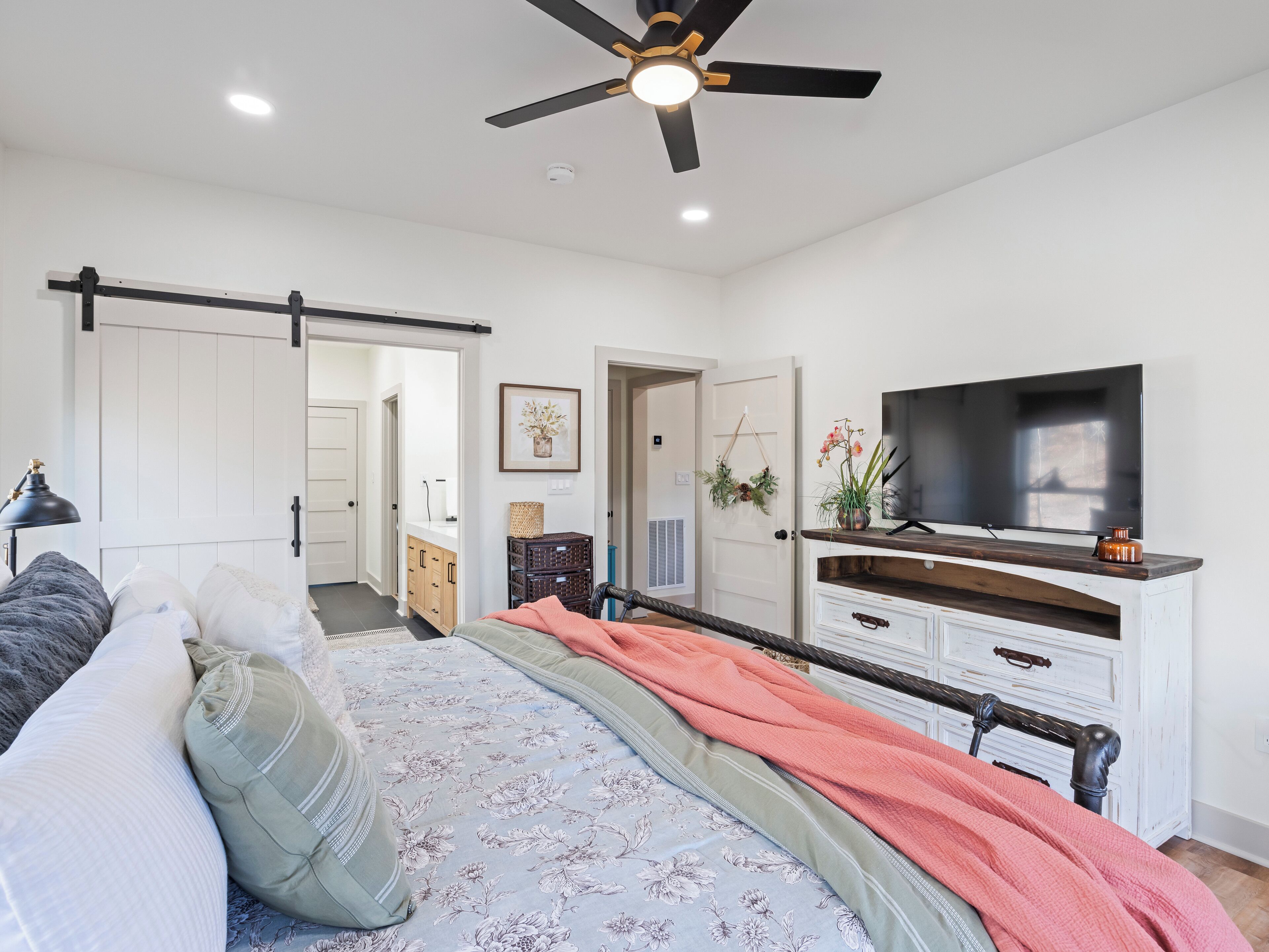 A bedroom with a bed, a TV, a ceiling fan, and a door leading to another room.