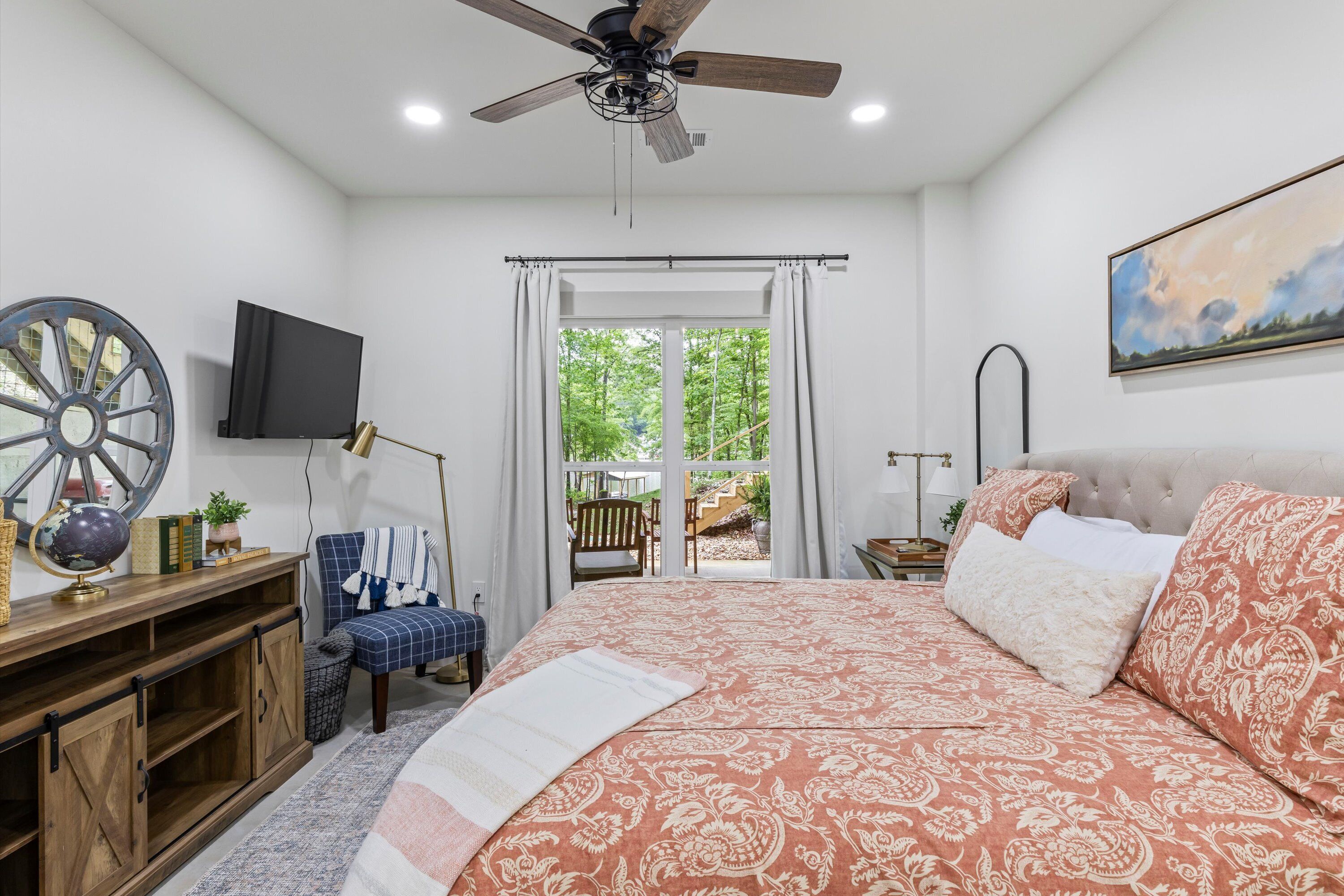 A bedroom with a large bed, a ceiling fan, a TV, a desk with a globe, a chair, and a window with curtains.