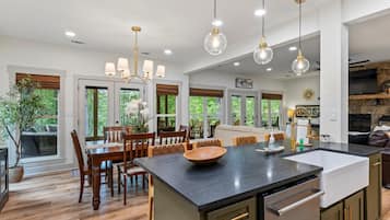 Une cuisine spacieuse dotée d’un grand îlot central, d’une table à manger en bois et de chaises. On y trouve des luminaires suspendus, un four encastré et une cheminée en pierre.