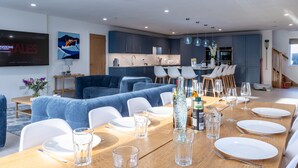 A modern dining area with a wooden table set for a meal, blue sofas, and a kitchen in the background. - The Old Pottery: Spacious 6‑Bed Beach House Sleeping 12 in Rhosneigr | Managed by Anglesey Holiday L (Rhosneigr)