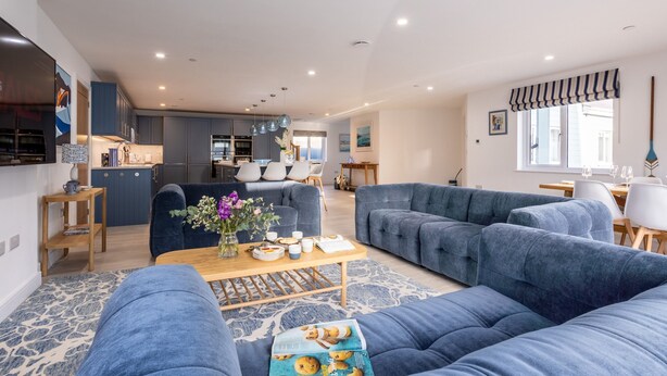 A modern living room with a large blue sofa, a wooden coffee table with a magazine, and a kitchen in the background. - The Old Pottery: Spacious 6‑Bed Beach House Sleeping 12 in Rhosneigr | Managed by Anglesey Holiday L (Rhosneigr)