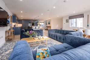 A modern living room with a large blue sofa, a wooden coffee table with a magazine, and a kitchen in the background. - The Old Pottery: Spacious 6‑Bed Beach House Sleeping 12 in Rhosneigr | Managed by Anglesey Holiday L (Rhosneigr)