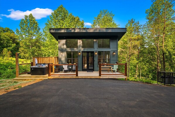 A modern two-story building with a deck, outdoor seating, and a view of the surrounding forest.