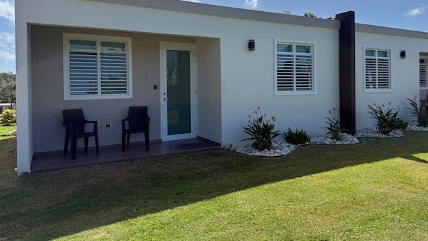 Uma casa moderna de dois andares com varanda coberta, duas cadeiras e um pequeno jardim com plantas e pedras.