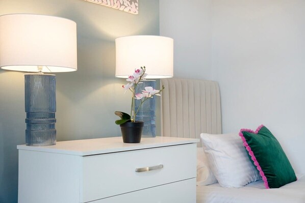 A bedroom with a white bedside table, two lamps, a potted orchid, a green pillow, and a white headboard.