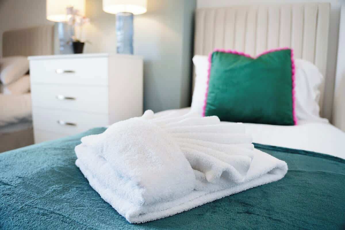 Un lit bien fait, recouvert d’une couvre-lit verte, avec un oreiller blanc bordé de vert et une table de chevet blanche dotée de tiroirs.
