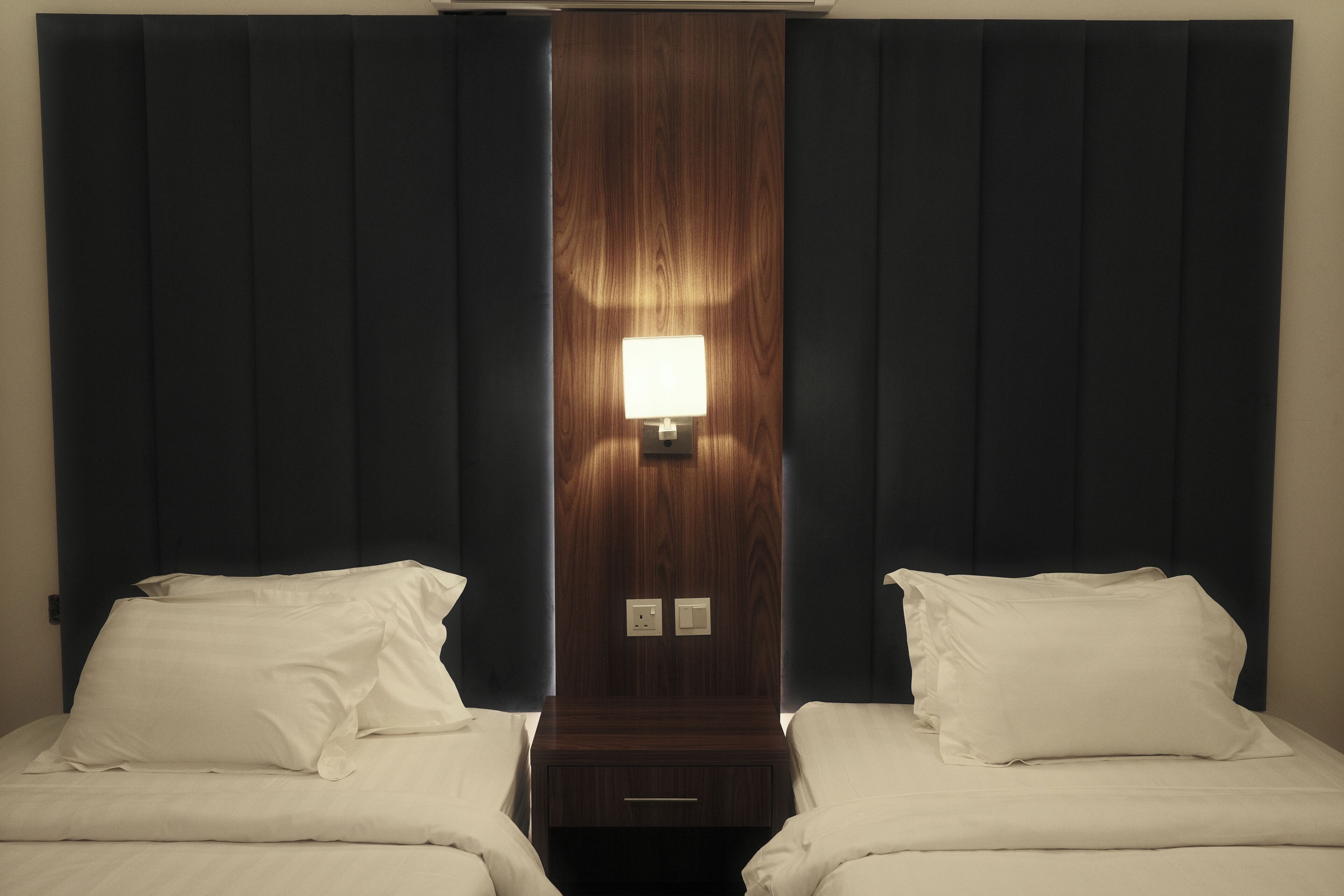Habitació d'hotel amb dos llits, un panell de capçalera de fusta, una tauleta de nit i una làmpada penjada a la paret.