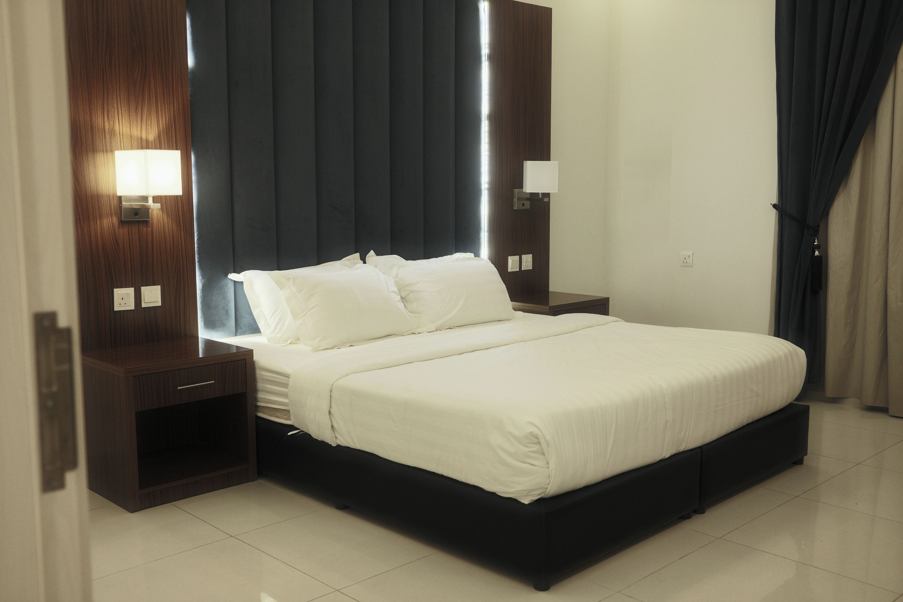 Habitación de hotel moderna con una cama grande, cabecero de madera oscura, mesitas de noche con lámparas y un amplio ventanal con cortinas.