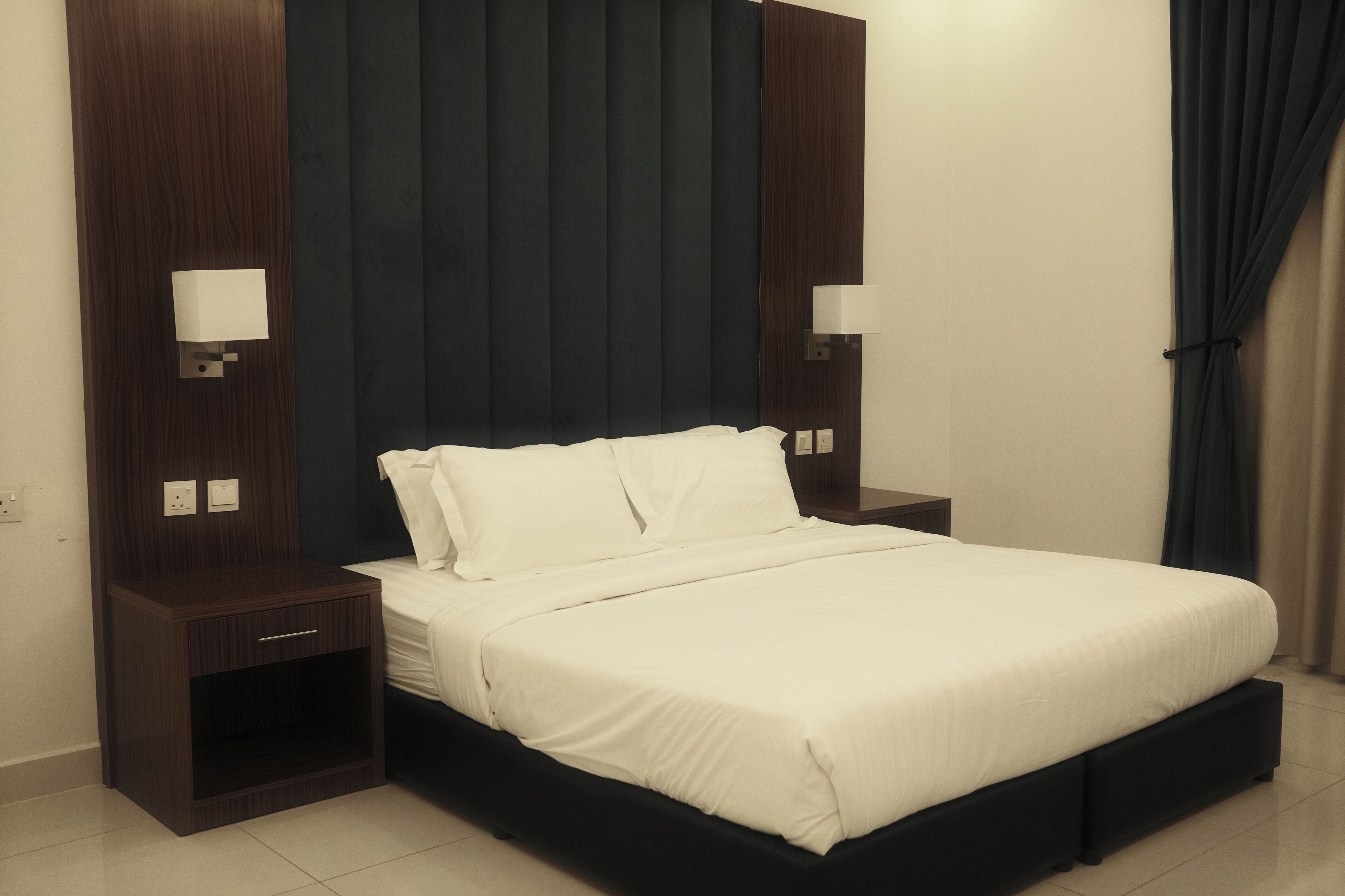 Ein modernes Hotelzimmer mit einem großen Bett, einem dunklen Holz-Kopfteil, Nachttischen mit Lampen und einem schweren Vorhang.