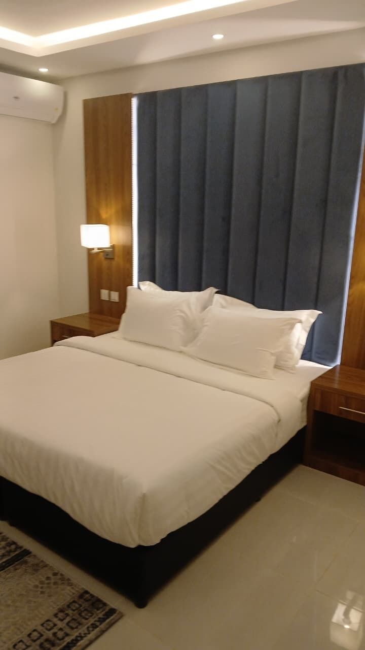 A hotel room with a bed, nightstands, a wall-mounted lamp, and a large window with curtains.