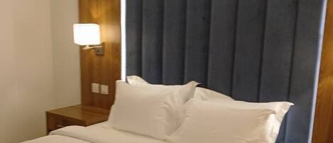 Habitación de hotel con cama, mesitas de noche, lámpara de pared y un gran ventanal con cortinas.