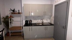 Une cuisine de taille réduite, équipée d’un évier, de placards et d’un plan de travail. Une porte se trouve à droite et un banc avec une plante à gauche.