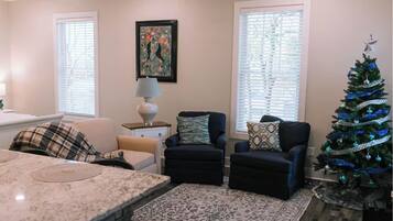 A living room with a marble coffee table, two armchairs, a small side table with a lamp, a framed artwork on the wall, and a decorated Christmas tree.