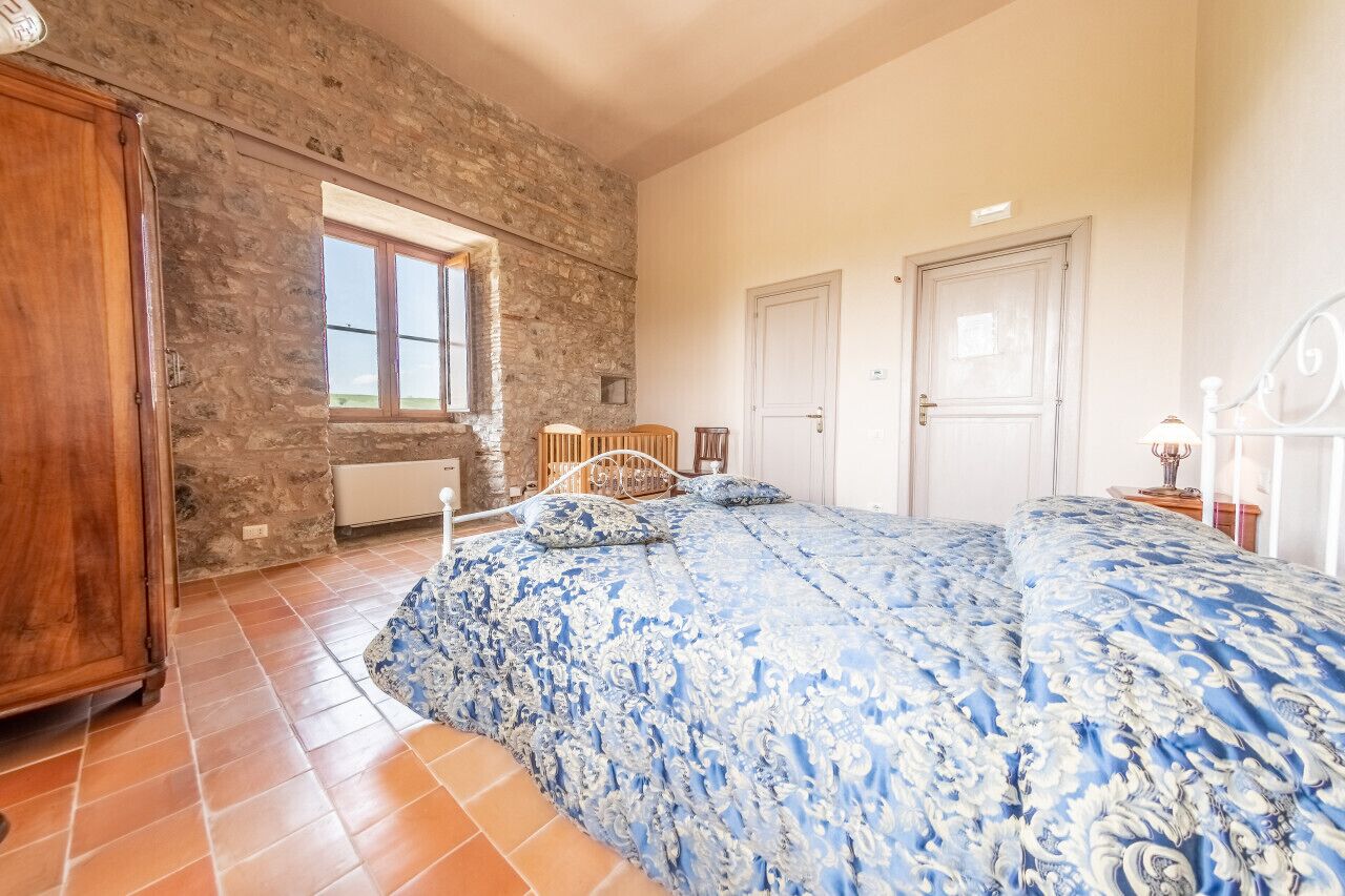A bedroom with a stone wall, a large bed with a blue floral bedspread, a wooden wardrobe, and a window with a view.