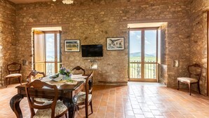 Un coin repas avec un mur en pierre, une table en bois et des chaises. Il y a deux fenêtres avec des rideaux, un téléviseur à écran plat fixé au mur et des tableaux encadrés au-dessus des fenêtres.